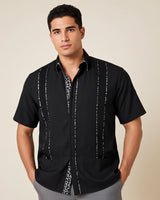 Man wearing a black guayabera shirt with white patterns on a beige background