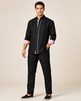 Man wearing a black guayabera shirt with pink cuffs and black pants on a beige background