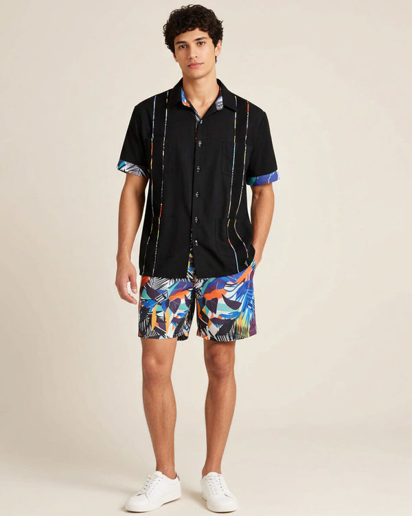 Man wearing a black guayabera shirt with colorful floral patterns and multicolored shorts on a beige background