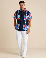 Man wearing a navy guayabera shirt with colorful floral patterns and white pants on a beige background