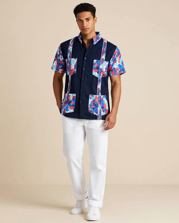 Man wearing a navy guayabera shirt with colorful floral patterns and white pants on a beige background