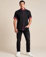 Man wearing a black guayabera shirt with colorful accents and black jeans on a beige background