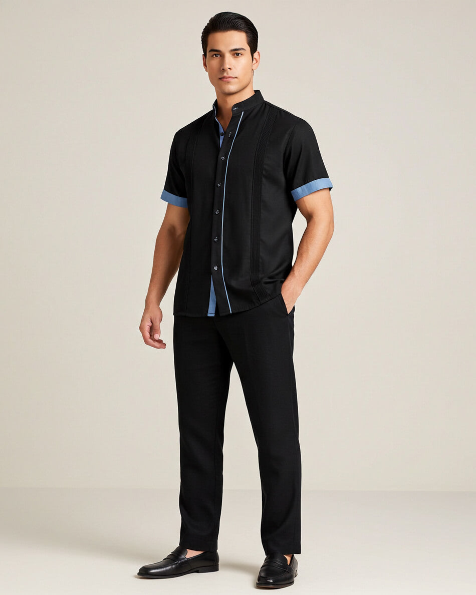 Man wearing a black short-sleeve guayabera shirt with blue accents and black pants on a plain background