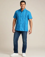 Man wearing a turquoise blue short-sleeve guayabera shirt and dark jeans on a beige background