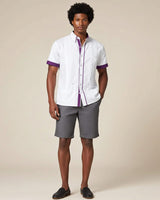 Man wearing a white guayabera shirt with purple accents and gray shorts on a beige background