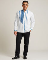 Man wearing a white embroidered guayabera shirt with blue patterns and black pants on a plain background