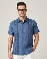 Man wearing a blue embroidered guayabera shirt with white pants on a plain background