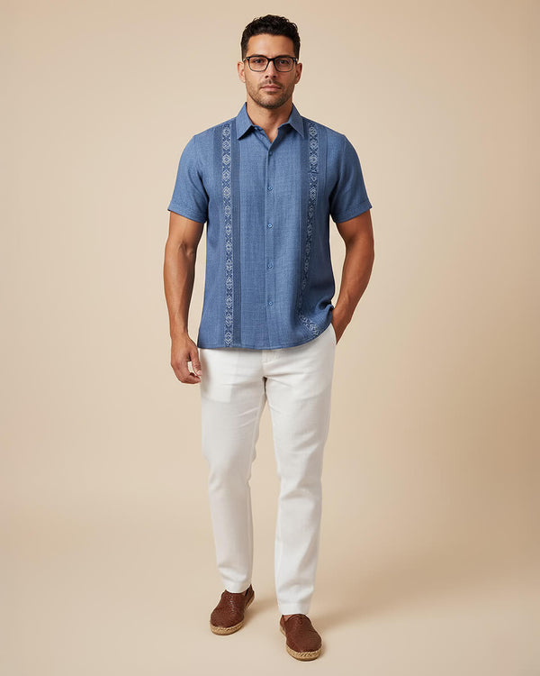 Man wearing a blue embroidered guayabera shirt with white pants on a beige background