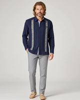 Man wearing a navy guayabera shirt with colorful patterns and gray pants on a white background