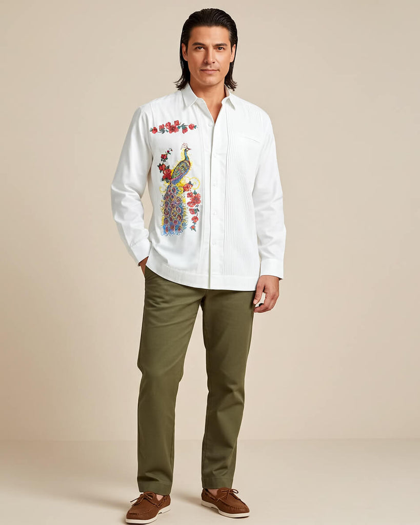 Man wearing a white embroidered guayabera peacock shirt with a colorful design, green pants, and brown shoes on a beige background.