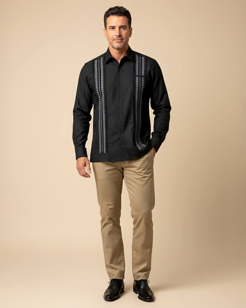 Man wearing a black guayabera shirt with white patterns and beige pants on a beige background